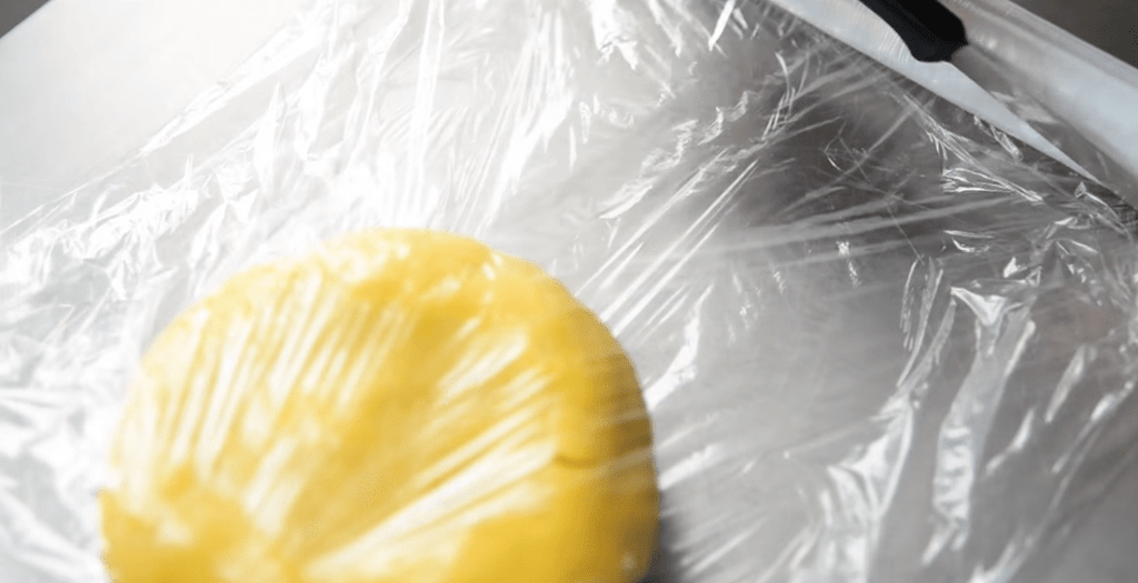 Ball of yellow cookie dough on plastic wrap on a metal work surface