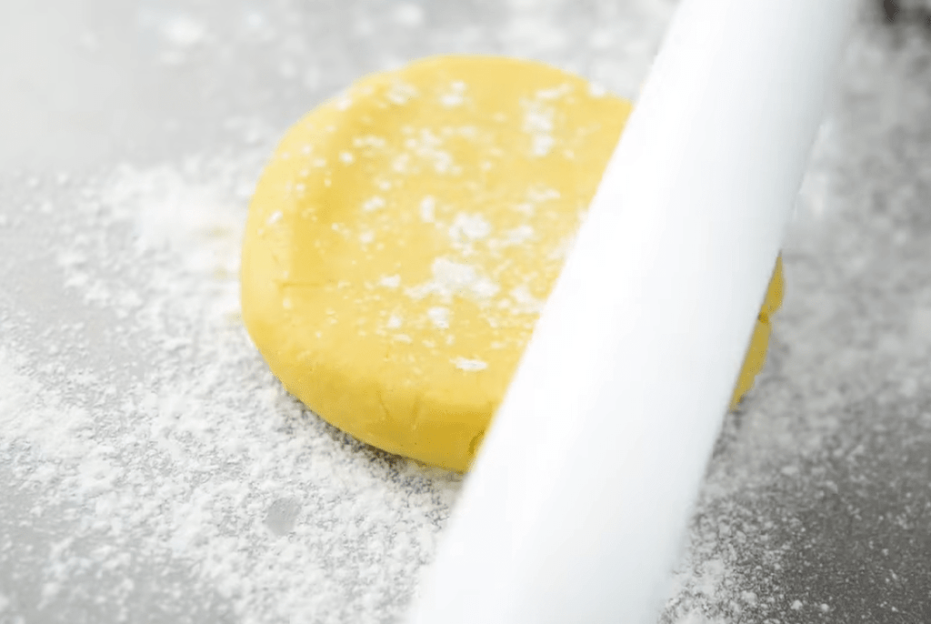 Rolling pin flattening yellow cookie dough on a lightly floured work surface