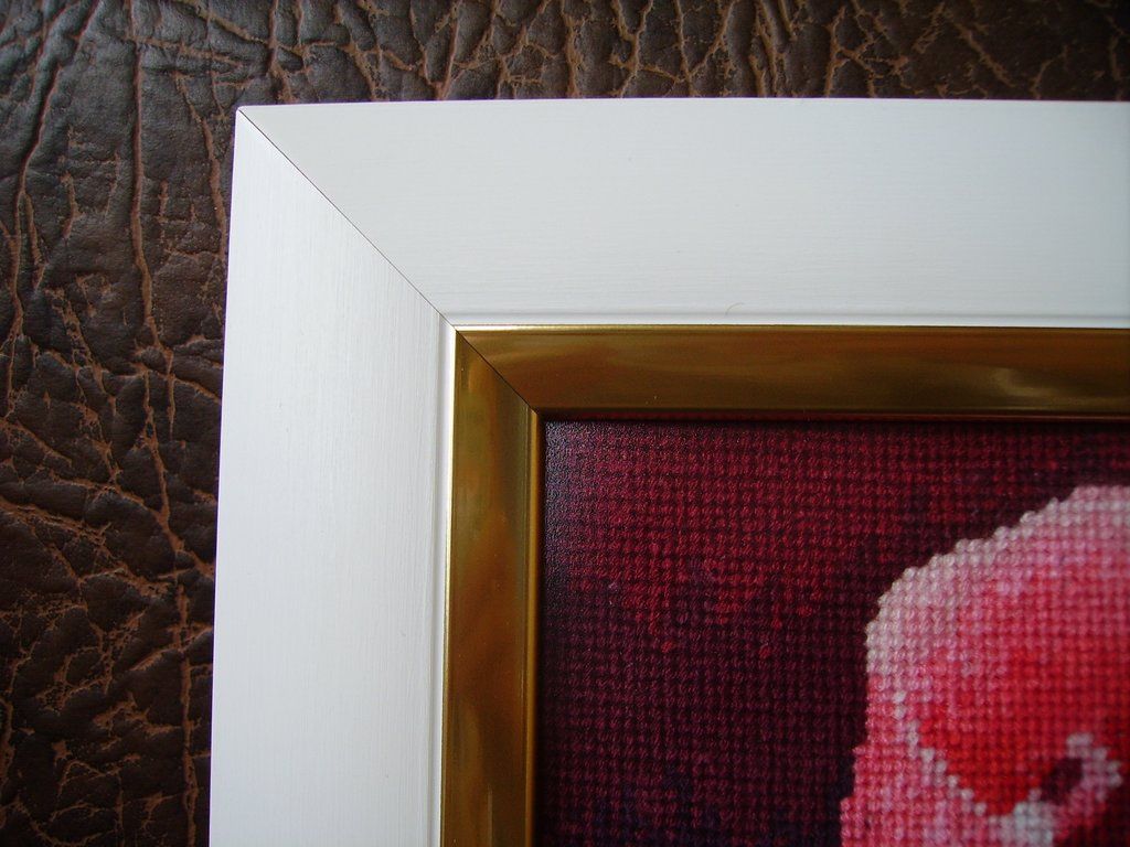 Close-up of a white-and-gold frame corner beside burgundy cross-stitch fabric and a pink rose petal