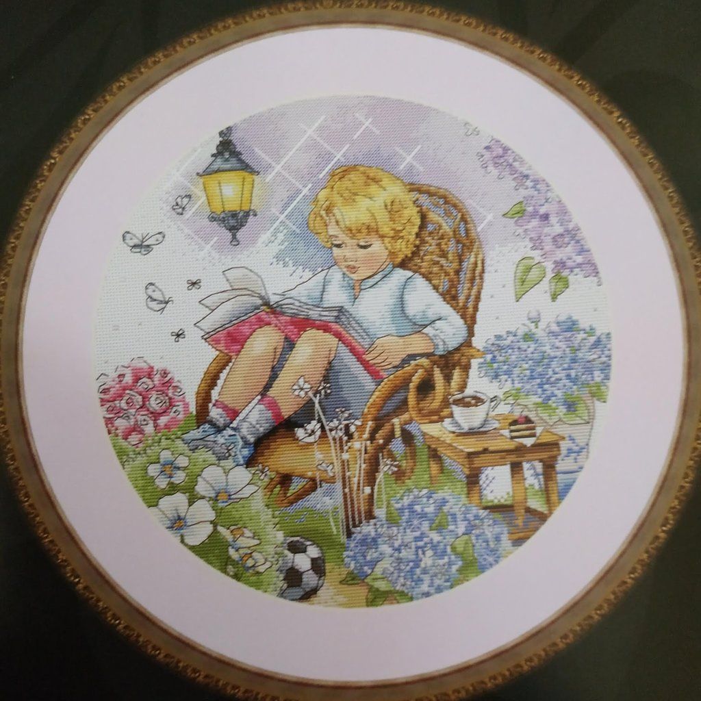 Framed circular cross-stitch portrait of a girl reading in a rocking chair among flowers