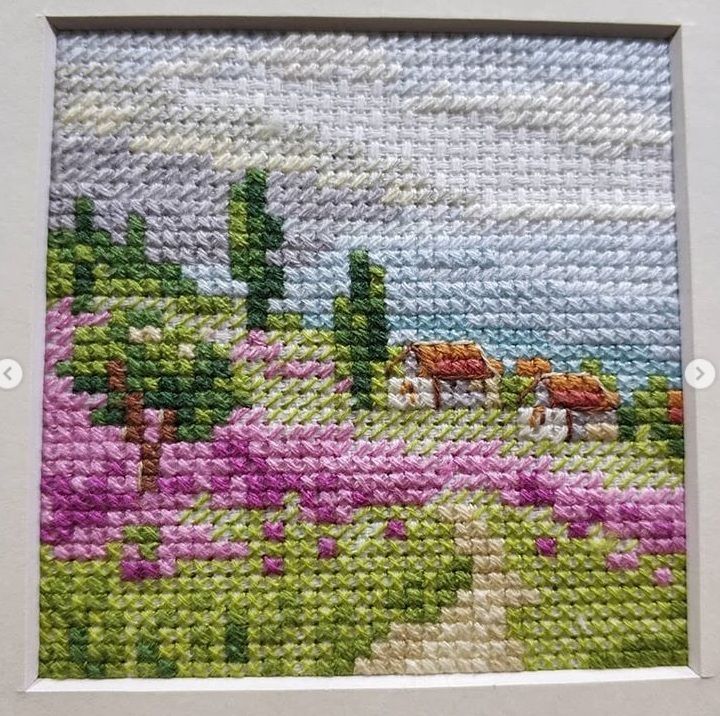 Miniature embroidery of a rural hillside with houses and pink flowers