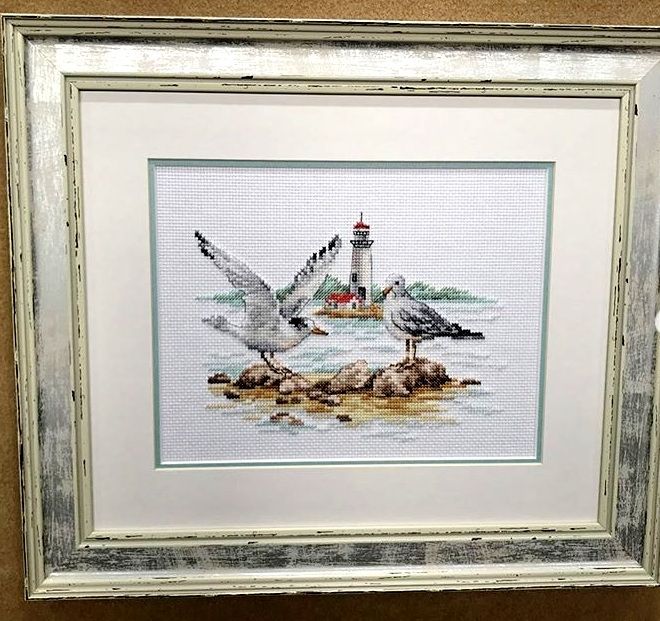 Framed embroidery of seagulls on rocks beside a lighthouse