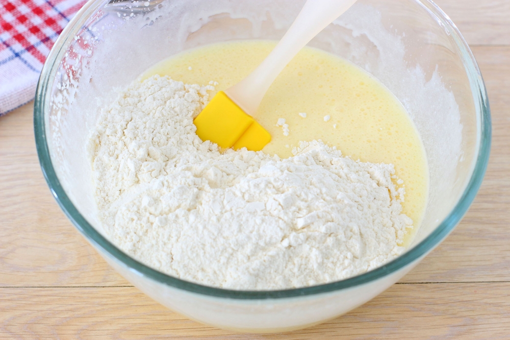 Flour being folded into pale muffin batter with a yellow silicone spatula